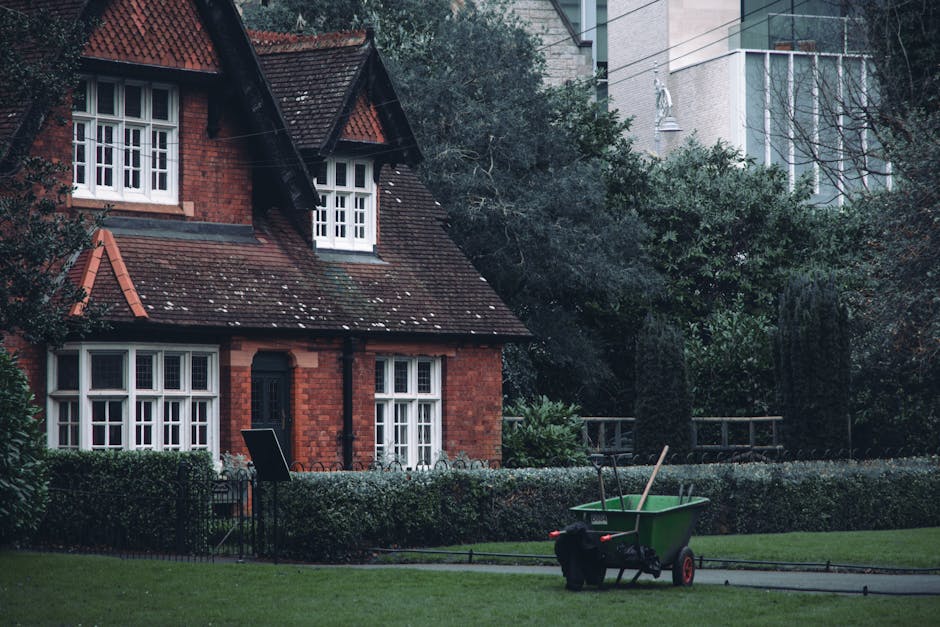 A traditional red-brick house with a steep, tiled roof and white-framed sash windows situated in a well-maintained garden. The garden features neatly trimmed hedges bordering a lush, green lawn, with 